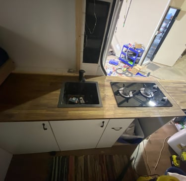 Custom camper van kitchen with wood countertop, black sink, and two-burner gas stove.