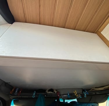 Custom white overhead storage cabinet inside a camper van with a wood paneled ceiling and storage line.