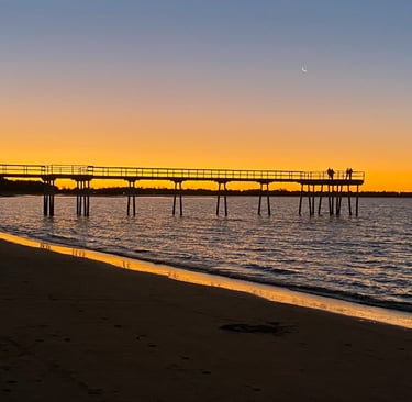Sunset at the beach with a silhouetted wooden pier and crescent moon over the ocean horizon.