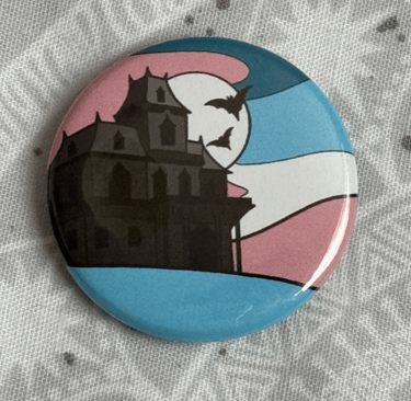 An image of a pin badge with a haunted house illustration in the colours of the trans flag.