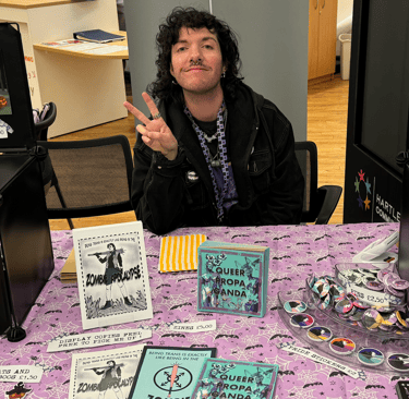 A photograph of the artist, an androgynous person with curly hair, they are doing the peace sign.