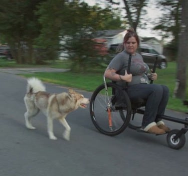 A white woman uses an off-roading wheelchair. She rushes down the road with a husky dog by her side.