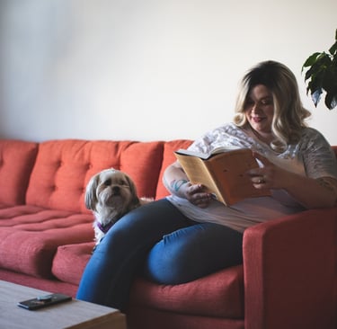 Women sat on a sofa with a dog reading a book.