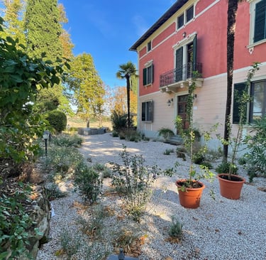 Villa Violetta with a red facade and gravel courtyard surrounded by lush Mediterranean plants.