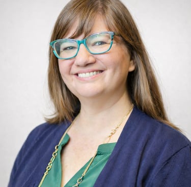 A smiling woman with brown hair and turquoise glasses wearing a navy cardigan and gold necklace.
