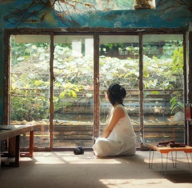 Person in a white dress sitting on the floor, peacefully looking out a large window into a lush gard