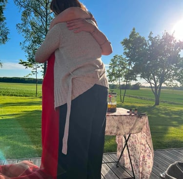 Two friends hugging on a sunny wooden deck overlooking a green field at sunset.