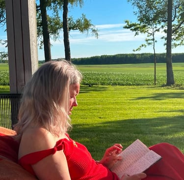 A woman in a red dress sits on a porch reading a book overlooking a green field and forest.