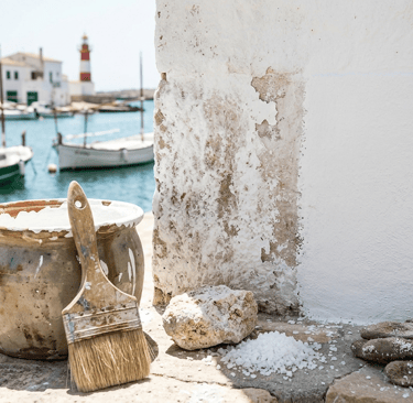A paintbrush and pot of white lime wash paint sit on a Mediterranean harbor wall near boats.