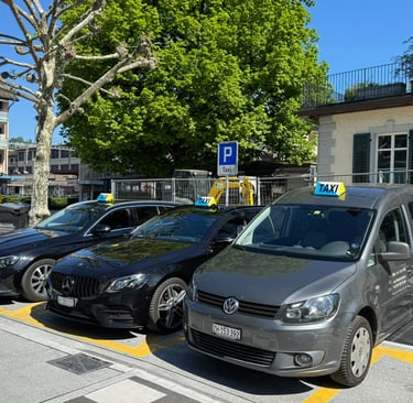 Three taxicars. One grey VW Caddy and two black Mercedes E-Classes parked on a swiss mainstation.