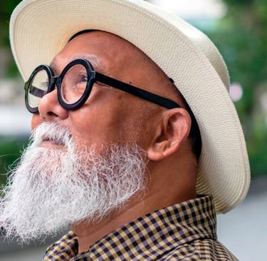 Stylish senior man with a white beard, round glasses, and a cream fedora hat looking up.