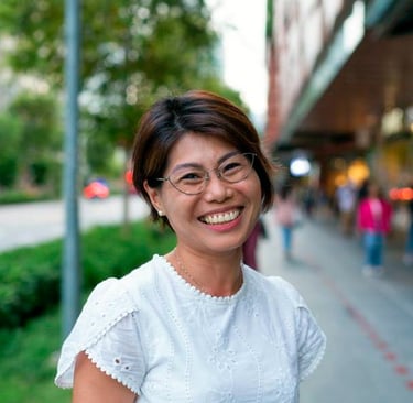 Smiling Asian woman with short hair and glasses wearing a white eyelet blouse on a city street.