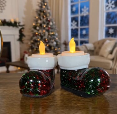 Two lighted Santa boot tea light candle holders on a table with a blurred Christmas tree background.