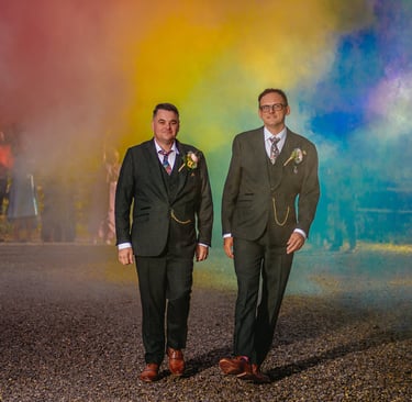 two grooms walking towards the camera with a rainbow of different coloured smoke bombs behind them