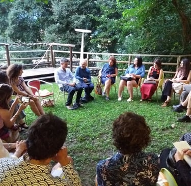 Cerchio di persone sedute all’aperto tra alberi e staccionata, in contesto informale