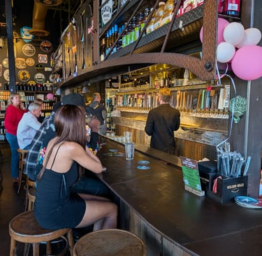 Customers sit at a rustic wooden bar in a craft brewery with many draft beer taps and industrial decor.