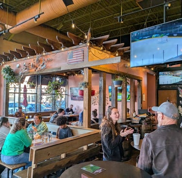 A cozy restaurant interior with customers dining at wood tables near sports TVs and hanging plants.
