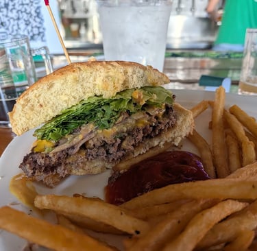 A gourmet cheeseburger cut in half served with crispy french fries and ketchup on a white plate at an outdoor bar.
