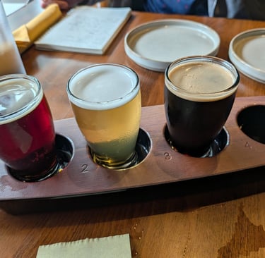 A craft beer flight with red, blonde, and dark stout samples on a wooden tasting paddle.