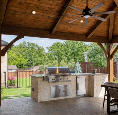 Covered outdoor kitchen in a North Texas backyard with a stone grill island, cedar patio cover, ceiling fan, and shaded seating area