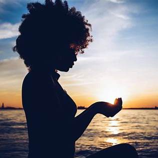 Black woman silhouette by the water at sunset, hands lifted as if holding the sun, calm, reflective