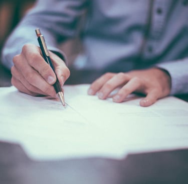 A person in a blue, button-up shirt sits at a desk writing on loose leaf paper