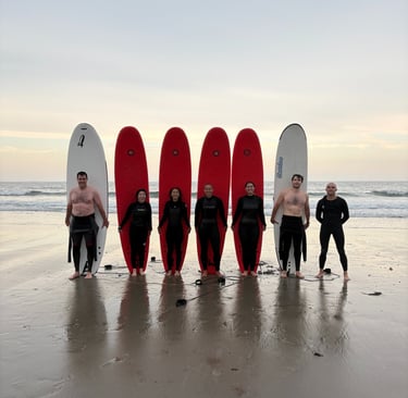 Surfers in wetsuits posing with red and white surfboards on a sandy beach at sunset.