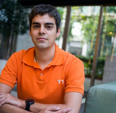 Um homem branco, Tiago Mitraud, veste uma camisa laranja e está sentado em uma cadeira verde.