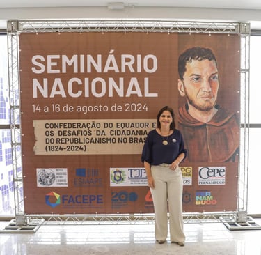 Priscila Krause, mulher branca, em pé na frente de um grande painel, vestindo blusa e calça.