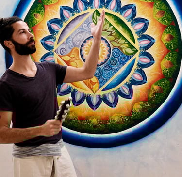 Bearded man in a yoga studio holding bells in front of a colorful sacred geometry mandala wall mural.