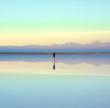 Person standing in a scene of mountains reflected in a lake. Photo by <a href="https://unsplash.com/