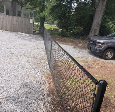 Black chain link fence separating a gravel driveway from a dirt path with trees and a grey pickup truck, enhancing security
