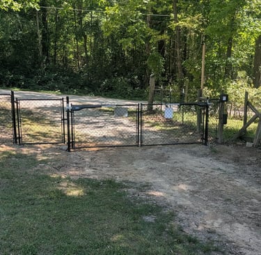 Automatic black chain link driveway gate with electric opener installed on a dirt road entrance.