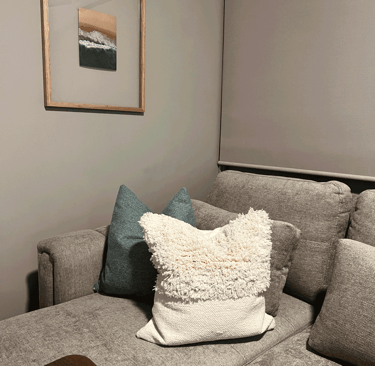 Rincón de sala con cojines de textura, cortinas enrollables y cuadro de paisaje costero | KARÄNA