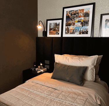 Cama individual con cabecera negra, cuadros y lámpara de lectura encendida | KARÄNA