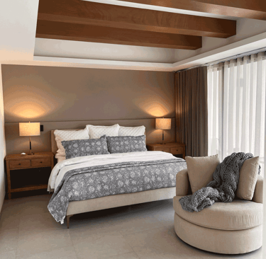 Recámara con acabados cálidos, tapiz neutro, cama con textiles grises y sillón individual | KARÄNA