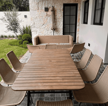 Comedor exterior con sillas y mesa de mimbre, junto a muro de piedra y jardín bien cuidado | KARÄNA