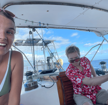 A smiling woman and a man in a red floral shirt steering a sailboat on the open ocean.