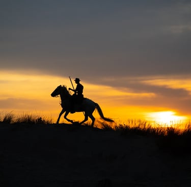 Silhouette of a cowboy riding a horse across sand dunes against a vibrant orange sunset sky.