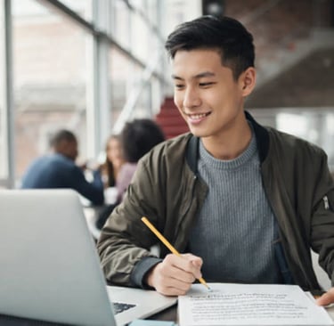 A smiling male student studying on a laptop and taking notes in a modern university library.