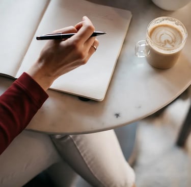 A person writing in a blank journal with a pen next to a latte at a cafe table.