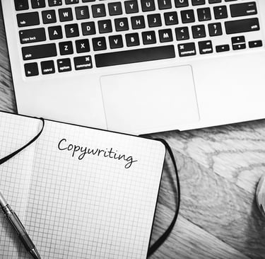 Black and white flat lay of a laptop, coffee, and copywriting notebook on a wooden desk.