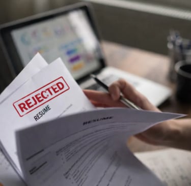 A job applicant holding a resume with a red REJECTED stamp on a wooden desk with a laptop.