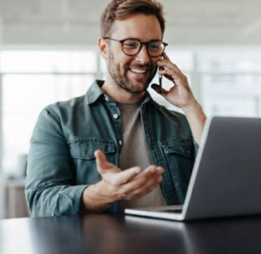 Smiling businessman wearing glasses talking on a smartphone while working on a laptop in a bright office.