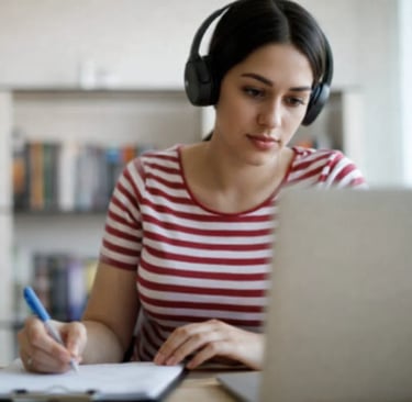 Young woman wearing headphones studying on a laptop and taking notes at a home office desk.