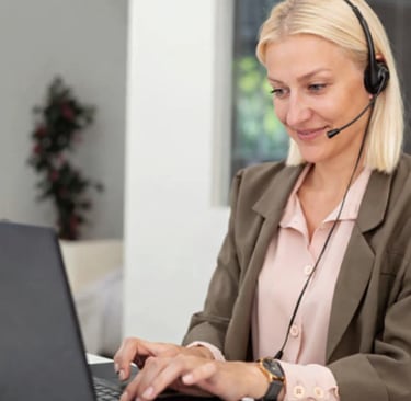 Blonde female customer support representative wearing a headset and working on a laptop in an office.