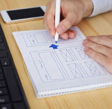 A web designer drawing a UI wireframe sketch on paper at a wooden desk with a computer keyboard.