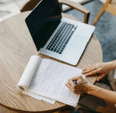 A copywriter taking notes on paper next to an open laptop on a wooden table.