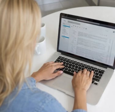 Blonde woman copywriter typing an email on a laptop at a white desk in a bright office.