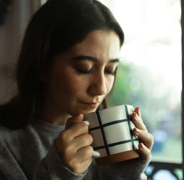 A young woman in a grey sweater holds a grid pattern coffee mug while enjoying a morning drink.